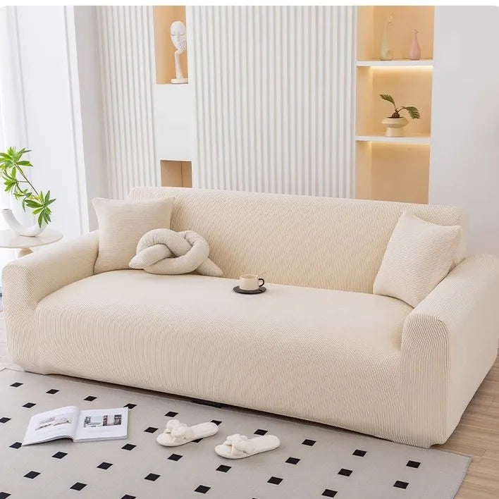 A beige stretchable full-wrap sofa slipcover displayed in a bright, modern living room setting.