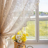 A sheer beige lace curtain with a beaded phoenix tail design hangs in front of a bright window overlooking green fields.