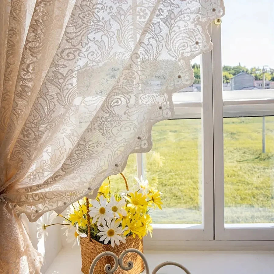 A sheer beige lace curtain with a beaded phoenix tail design hangs in front of a bright window overlooking green fields.