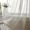 A sheer white curtain hangs in a window, draped elegantly with a rod pocket, next to a white vase of flowers.