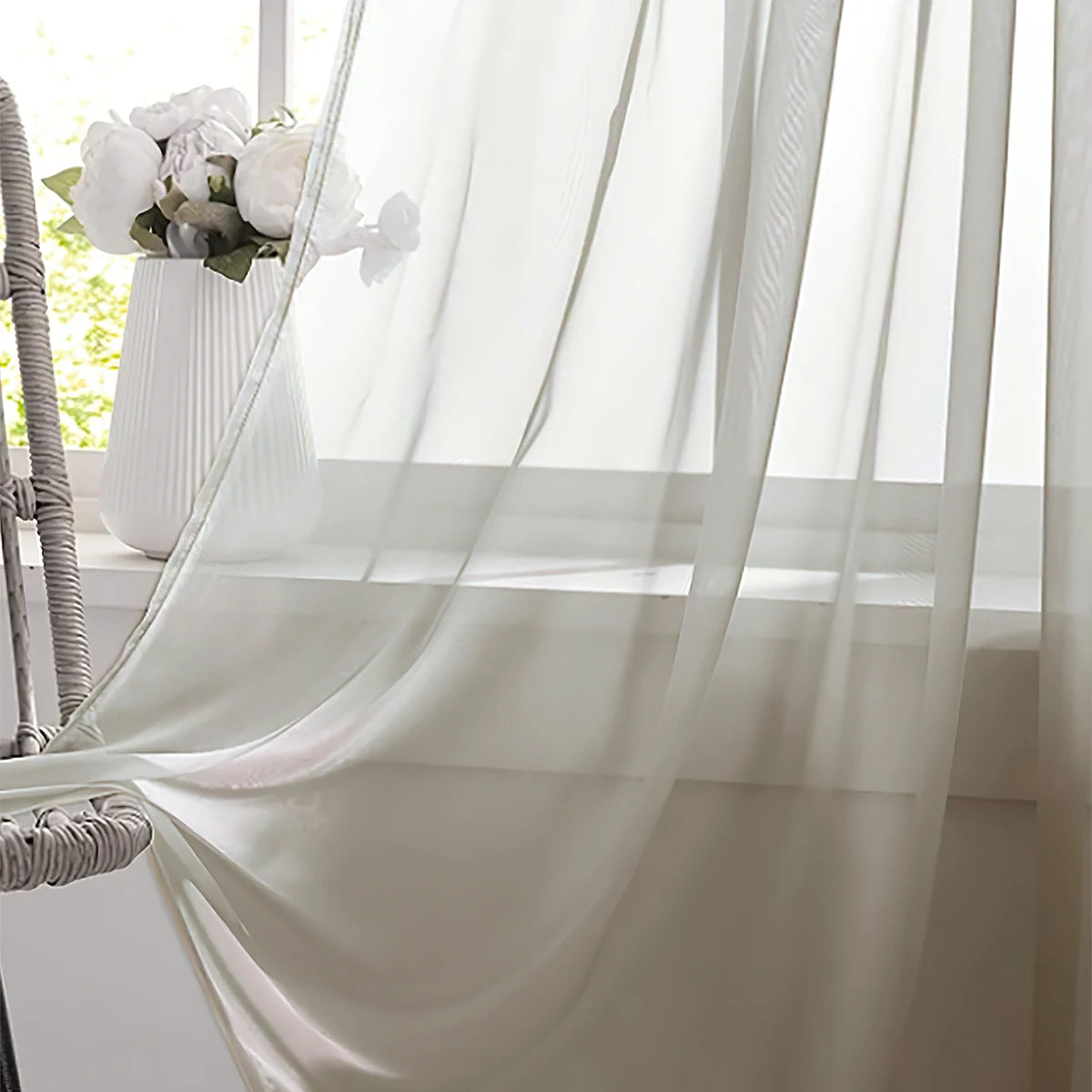 A sheer white curtain hangs in a window, draped elegantly with a rod pocket, next to a white vase of flowers.