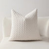 A white, textured decorative pillow cover with a zipper closure resting on a light-colored sofa.