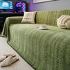 A green chenille sofa protector with a fishbone pattern is displayed on a bed, next to a pillow and a cup.
