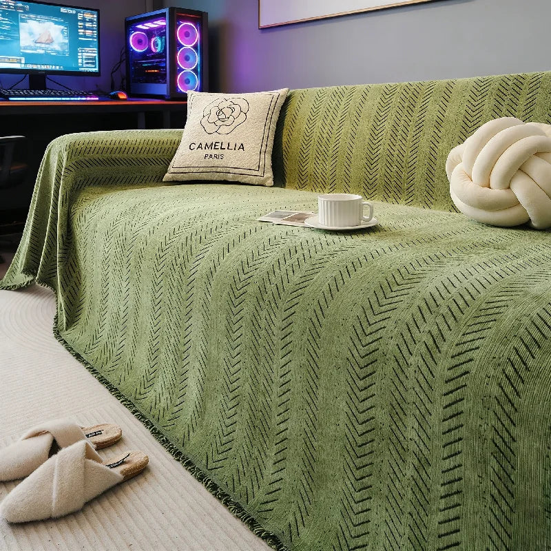 A green chenille sofa protector with a fishbone pattern is displayed on a bed, next to a pillow and a cup.