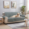 A beige reversible sofa slipcover in a sage green color is displayed on a light wood floor with modern wall art and plants.
