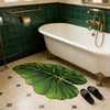 A large green leaf-shaped bathroom mat with a rubber backing placed on a tiled floor next to a white bathtub.