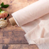 A rolled pure French linen bed sheet in a pale beige color resting on a wooden surface next to a pink rose.