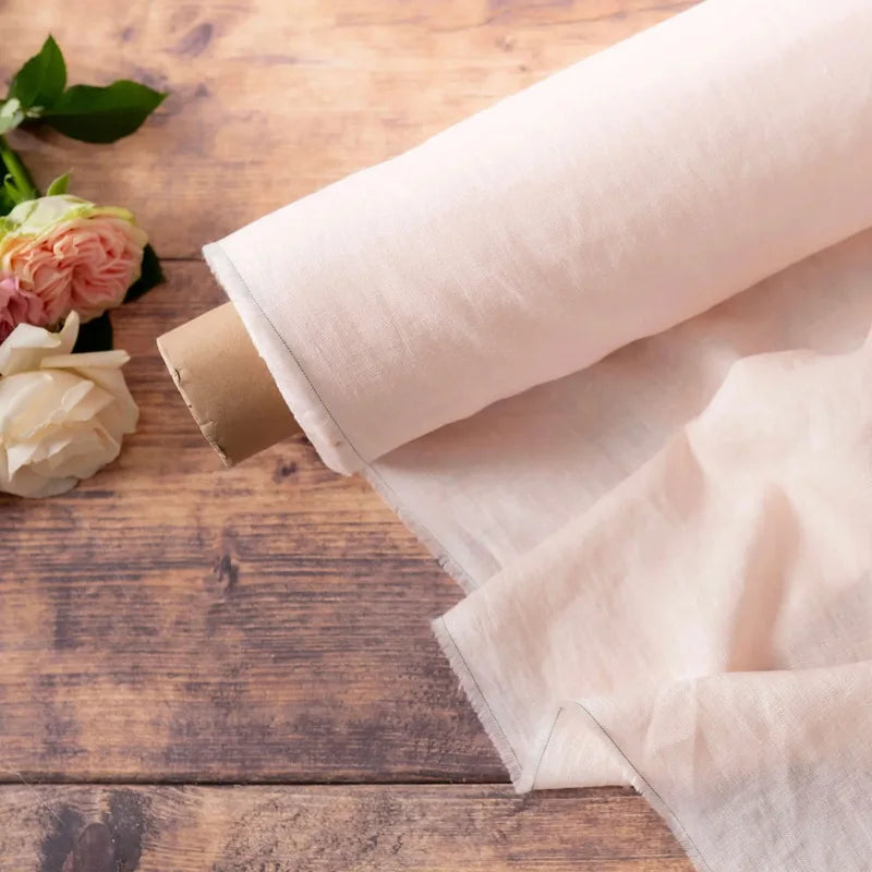 A rolled pure French linen bed sheet in a pale beige color resting on a wooden surface next to a pink rose.