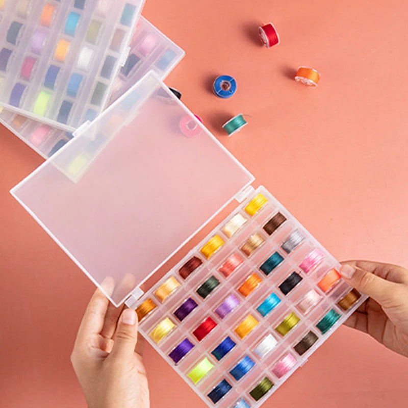 A hand holds open a clear plastic box containing a multicolor sewing bobbin set with various colored bobbins.