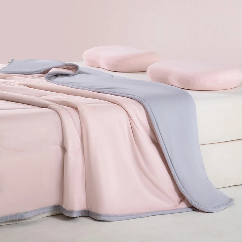 A pink and gray ice silk summer cooling blanket draped over a white surface with two pink pillows.