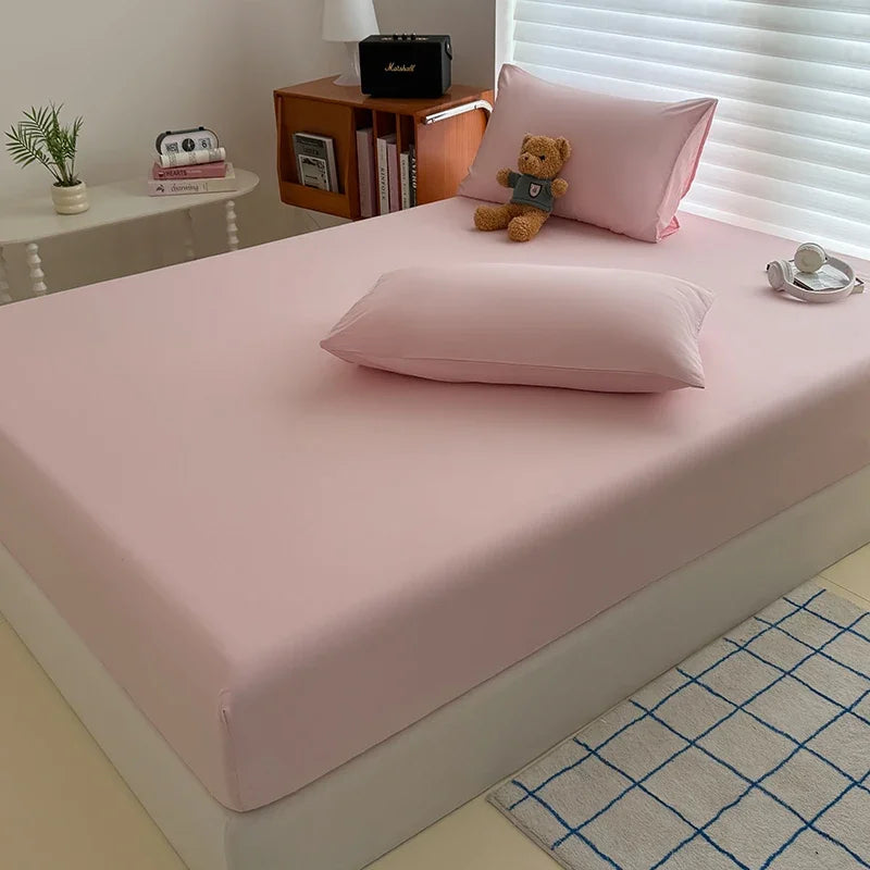 A pink fitted bed sheet cover is displayed on a bed, with pillows and a small black box nearby.