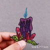 A hand holds an embroidered patch featuring a purple candle with a blue flame and red floral elements.