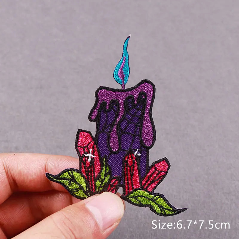 A hand holds an embroidered patch featuring a purple candle with a blue flame and red floral elements.