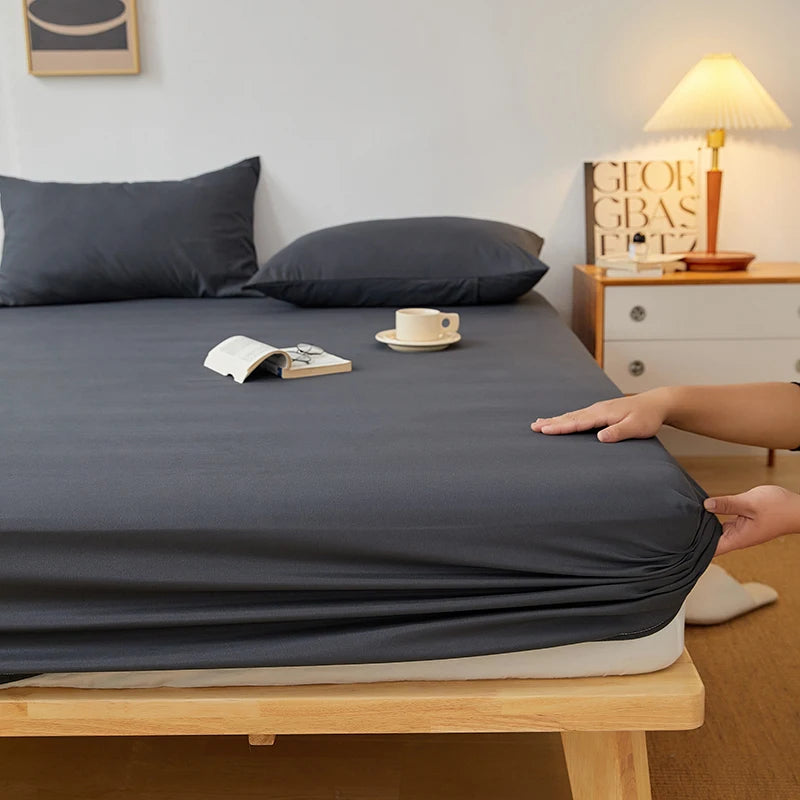 A dark gray fitted sheet is laid out on a wooden bed frame with a side table and lamp in the background.