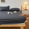 A dark gray fitted sheet is laid out on a wooden bed frame with a side table and lamp in the background.