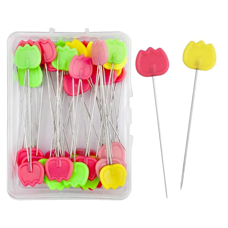 A clear plastic box containing numerous nickel-plated dressmaking pins with colorful heads in shades of green, yellow, and pi