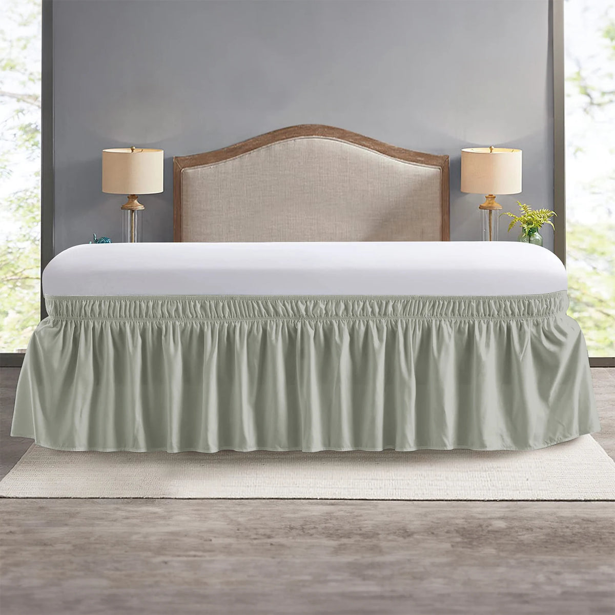 A light sage green wrap around elastic bed skirt covering a bed with a wooden headboard and bedside lamps.