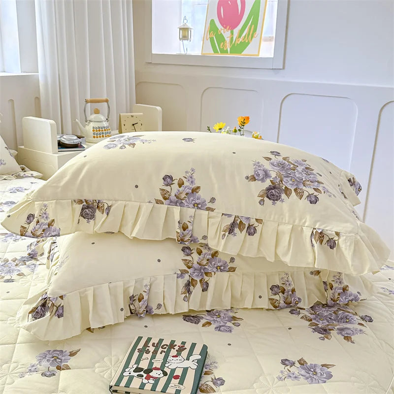 Two floral cotton pillow covers in pale yellow and purple are stacked on a quilted bed with a book.