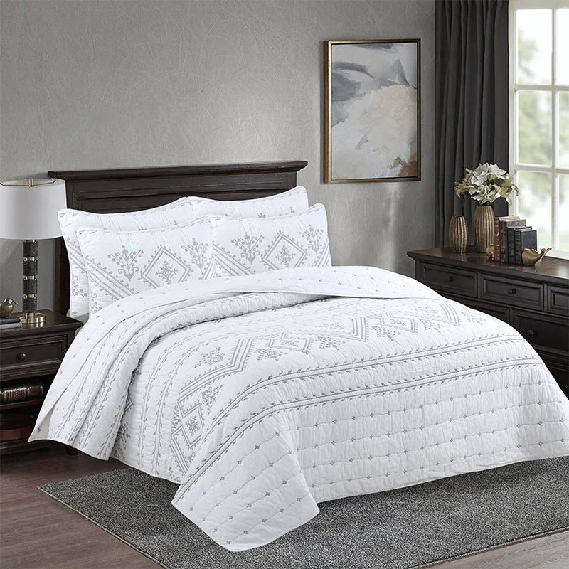 White embroidered bedding set with a quilted cover and patterned duvet on a dark wood bed.