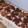 A close-up of a rustic farmhouse maple leaf table runner featuring various shades of brown and copper leaves on a white woode