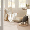 A beige linen textured throw pillow cover draped over a woven chair in a bright, airy room.