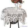 White floral lace tablecloth set with a vase of flowers and an hourglass on a dark table.