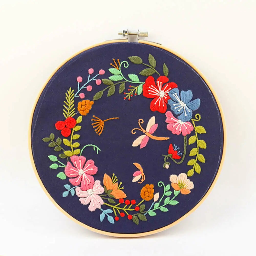 A circular embroidery hoop featuring a floral wreath of red, pink, blue, and green fabric stitches on a dark blue background.
