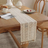 A cream vintage crochet lace table runner with fringe placed on a wooden table setting.
