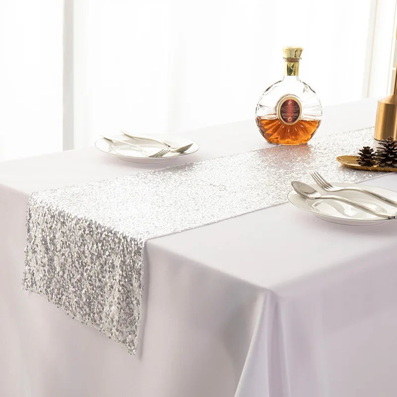A silver sequin table runner is displayed on a white tablecloth with glassware and silverware.