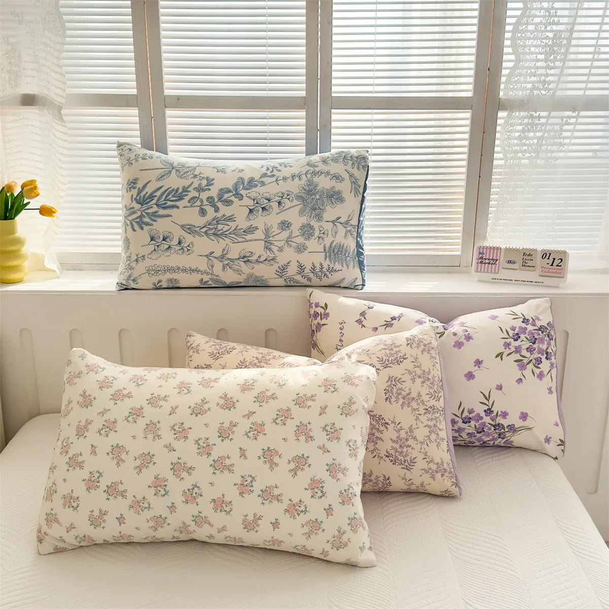 A collection of floral printed pillow covers in shades of blue, purple, and cream displayed on a white surface near a window.
