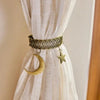 A close-up of a natural fiber curtain tieback featuring a woven rope clip with a gold star and a crescent moon charm.
