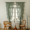 A pair of sheer sage green lace curtains hang in front of a white window with a wooden chair and table.