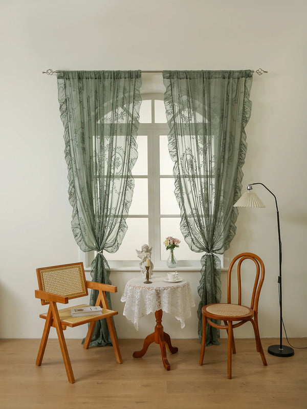 A pair of sheer sage green lace curtains hang in front of a white window with a wooden chair and table.