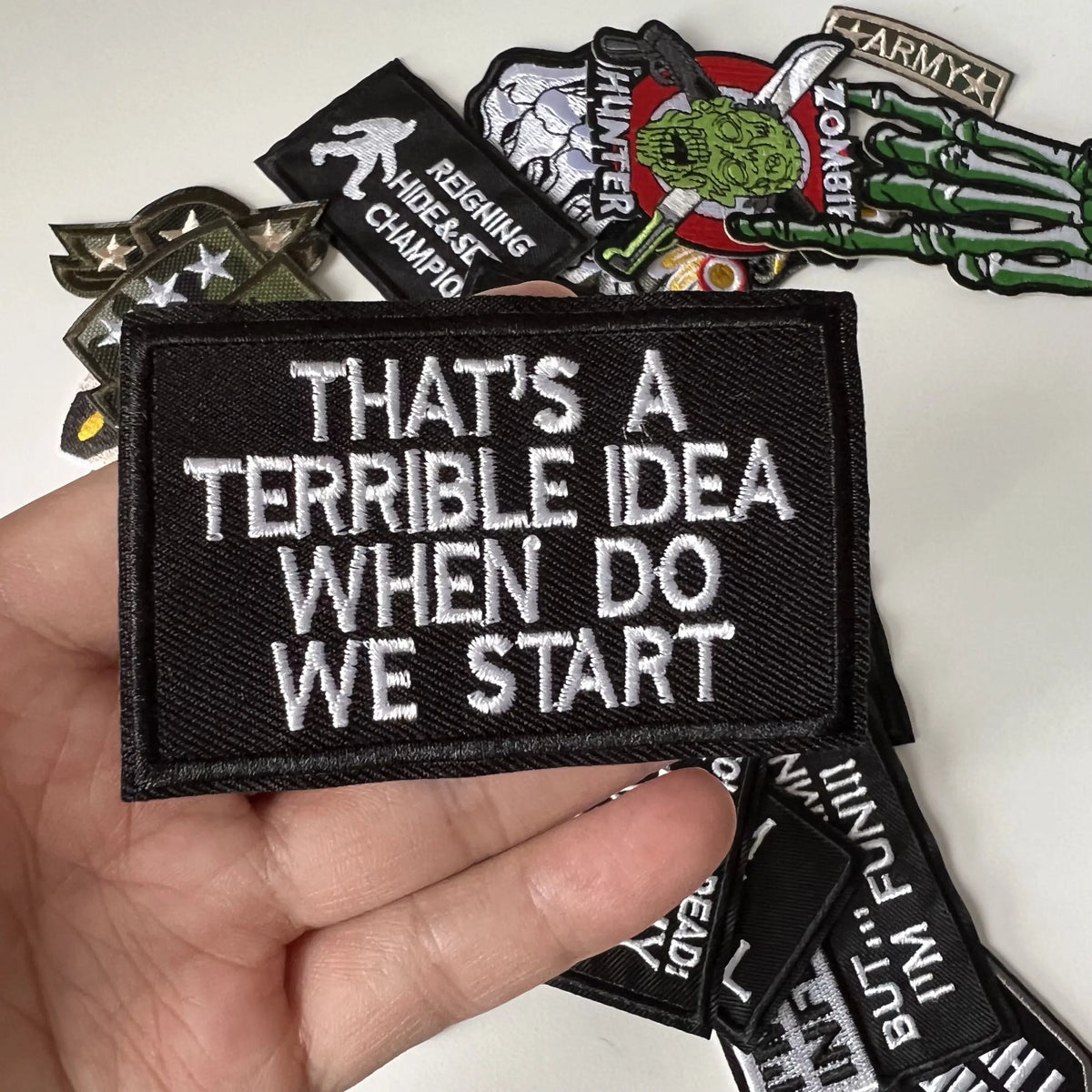 A hand holds several black iron-on slogan patches featuring text like That's a Terrible Idea When Do We Start