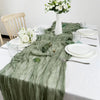 A long, sheer sage green gauze table runner draped elegantly on a white tablecloth with white plates and glassware.