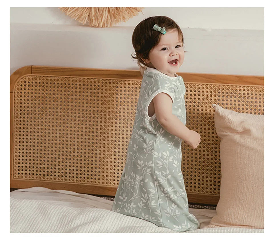 A smiling baby wearing a light green patterned soft cotton sleeping bag or swaddle, lying on a white surface.