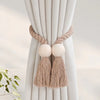 Hand-woven wooden curtain tiebacks with beige rope and fringed tassels against a light fabric backdrop