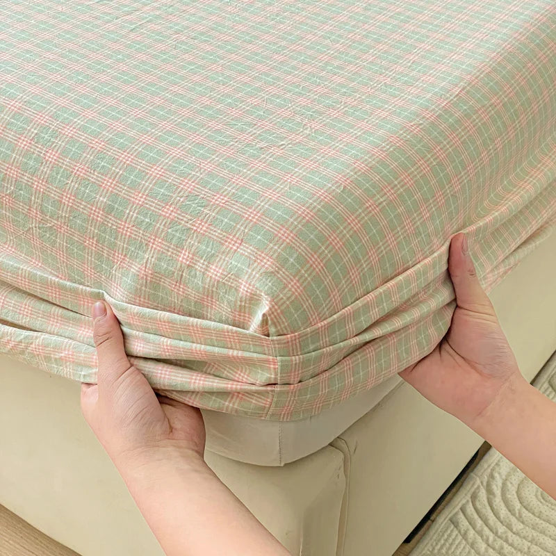 Hands are holding up a light green and pink plaid pure cotton fitted sheet.