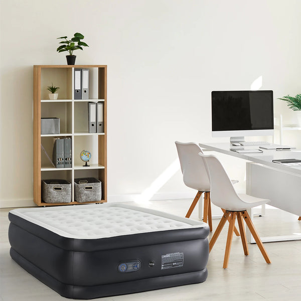 A black air mattress is placed on the floor next to a wooden shelf and a white desk.