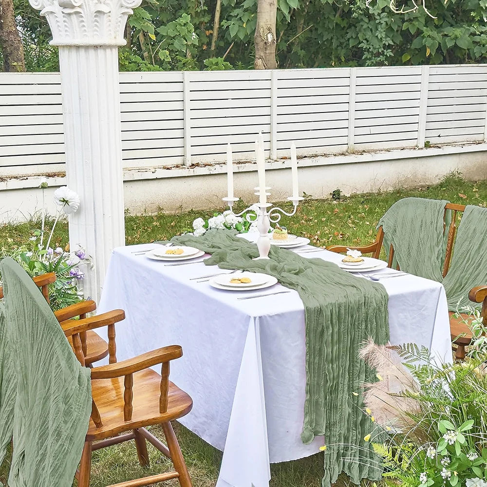 A white linen table set with a sage green cheesecloth runner and candles outdoors in a garden setting.