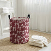 A round, maroon canvas laundry basket with a white bow pattern sits on a woven rug next to sheer white curtains.