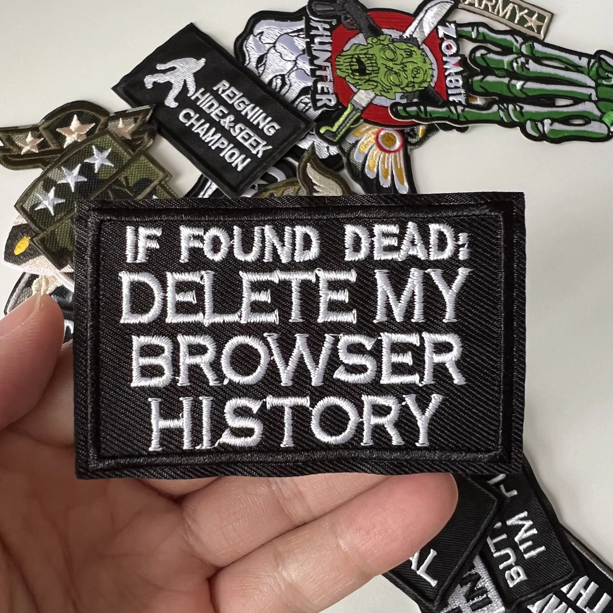 A hand holds several black iron-on slogan patches featuring text like If Found Dead Delete My Browser History and various mil