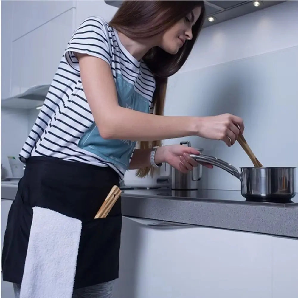 A woman wearing a black waist apron is cooking in a kitchen, holding a wooden spoon over a pot.
