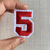 A hand holds a red and white embroidered number 5 patch for apparel.