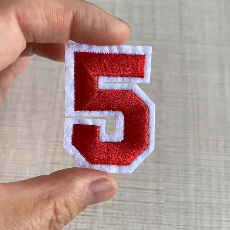 A hand holds a red and white embroidered number 5 patch for apparel.