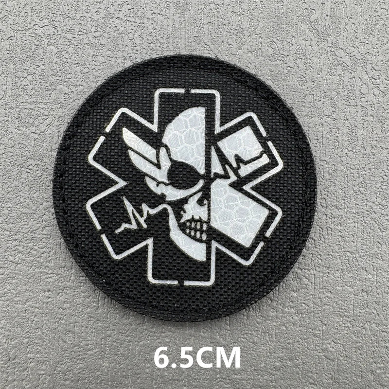 A black embroidered patch featuring a white medical cross design with an eye motif on a gray background.