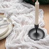 A white crinkled cheesecloth table runner draped over a wooden surface with a tall white candle and festive greenery.