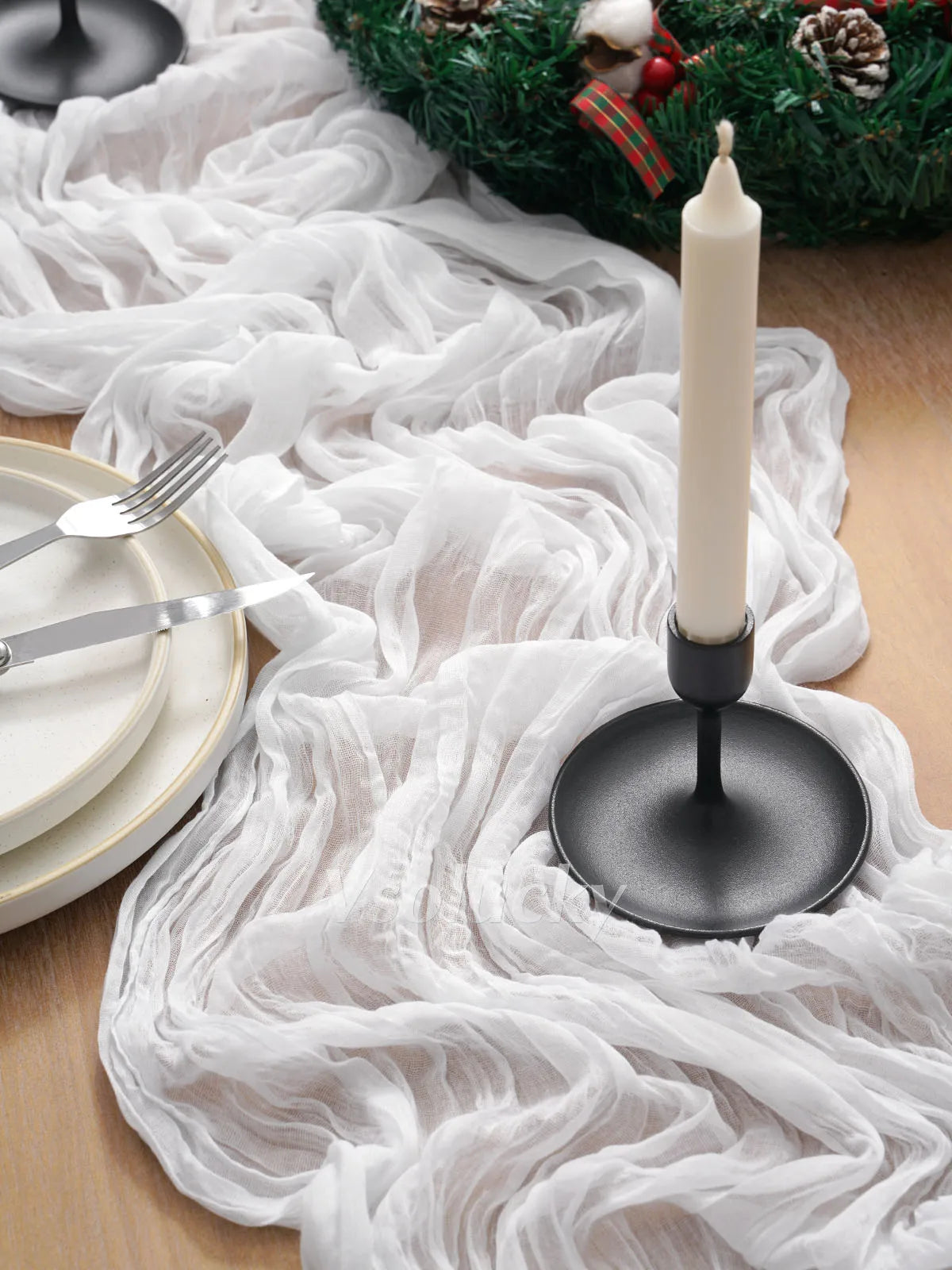 A white crinkled cheesecloth table runner draped over a wooden surface with a tall white candle and festive greenery.