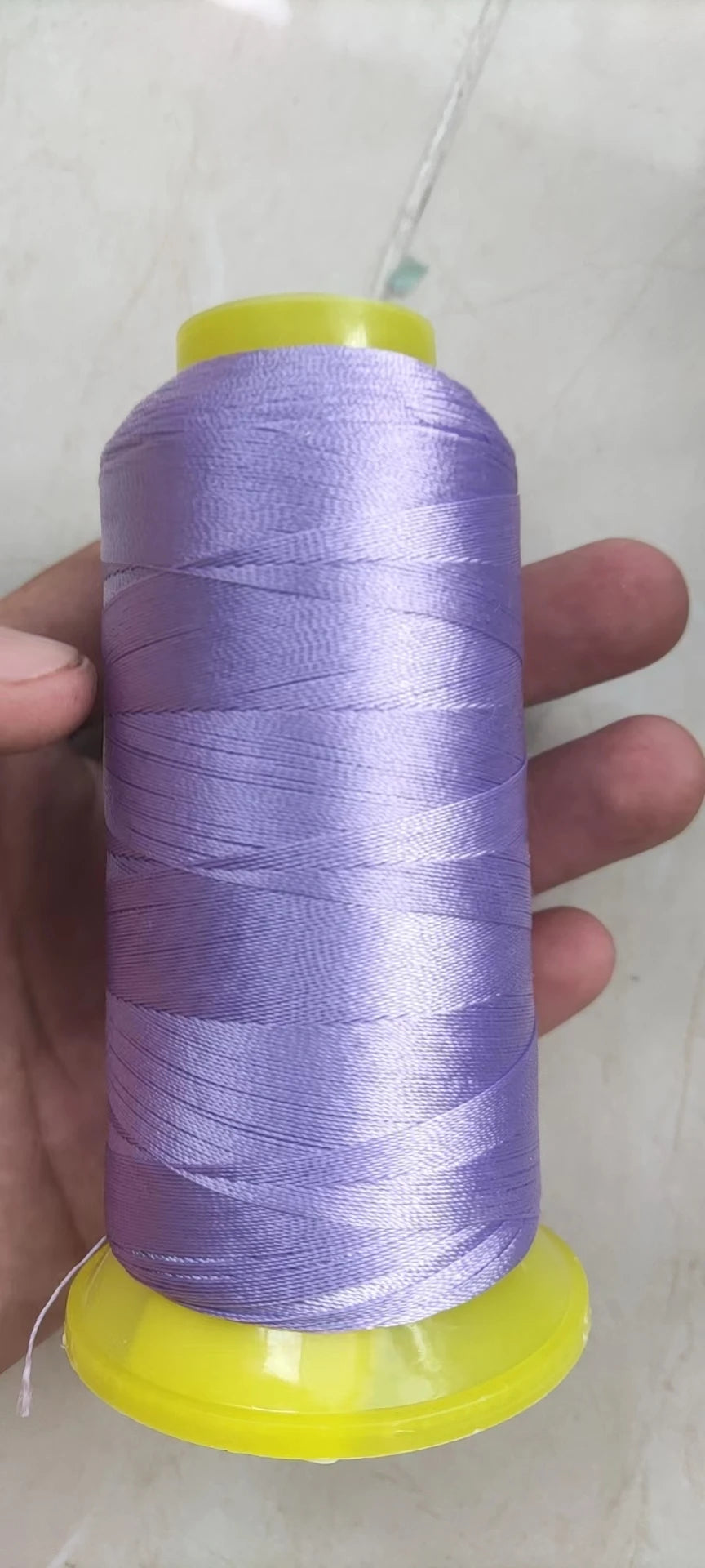 A spool of purple high tenacity polyester sewing thread with a yellow plastic core is held in a hand.