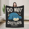 A black throw blanket featuring a cartoon blue cat face with the text DO NOT DISTURB I'M BUSY